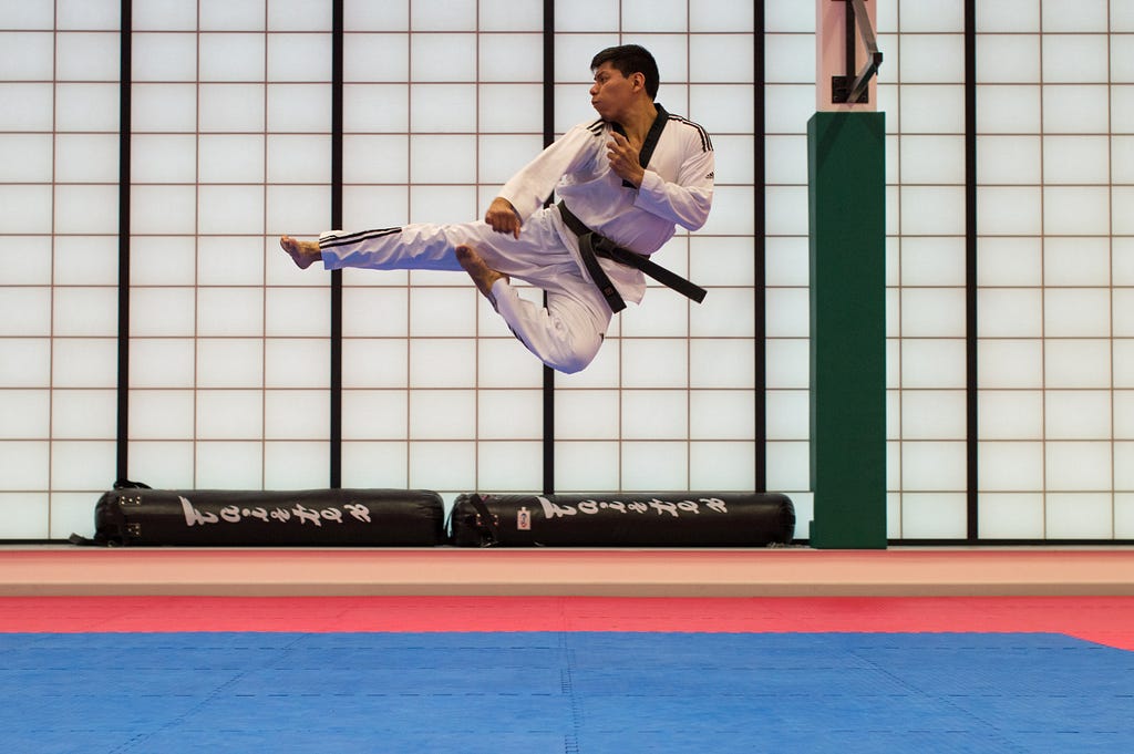 Man with a black belt in Tae Kwon Do performs jump side kick with right leg. He faces to right. Hands protect chest.