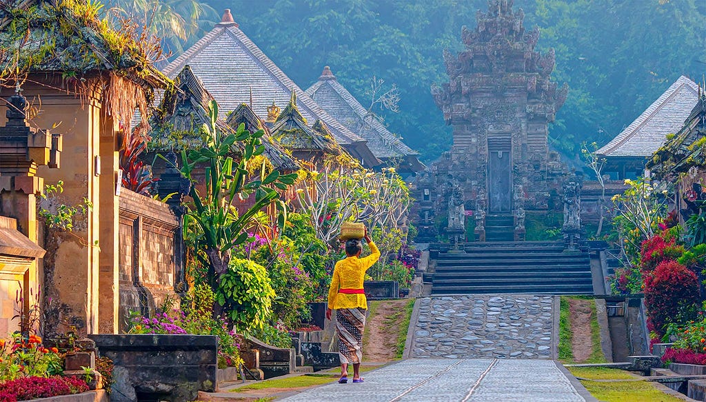 A Balinese woman in traditional attire carrying a basket on her head walks through the picturesque village of Penglipuran, Bali, under a hazy sky, with its lush greenery and distinctive architectural style of stone temples and gates in the background.