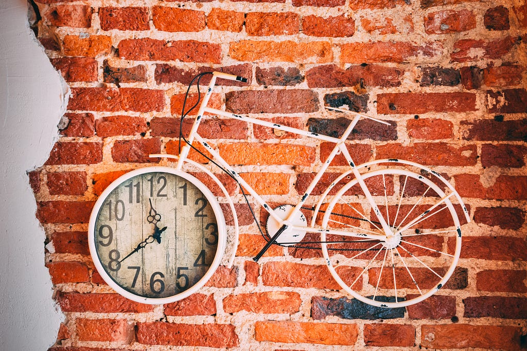 A bicycle with a clock in the front wheel against an orange brick wall.