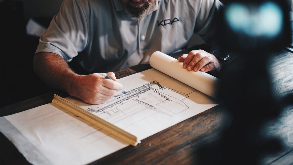 A male architect with a blue shirt sitting on a wooden desk with a constructional drawing in front of him.