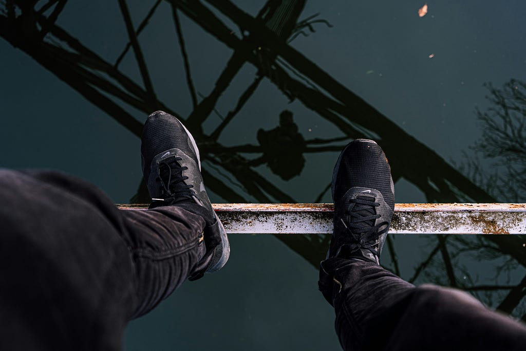 A point of view photo of a man’s feet, precariously standing on a metal beam, overlooking dark water.