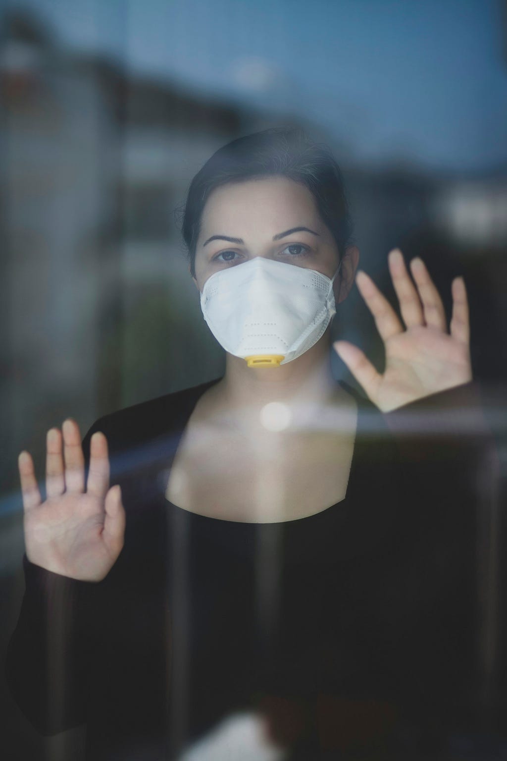 A woman wearing a mask looking out the window with her hands on the window