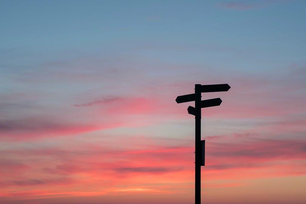 This is a photo of a signpost against a beautiful pink and orange sunset sky. The signpost is black and has three arms pointing in different directions. The sky is a gradient of pink and orange with wispy clouds. The photo is taken from a low angle, looking up at the signpost.