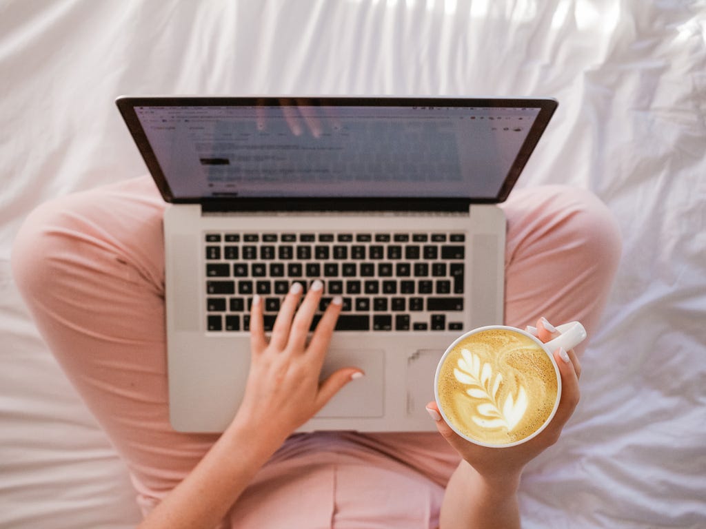 Woman sitting on her bed with her legs crossed drinking coffee and using her laptop.