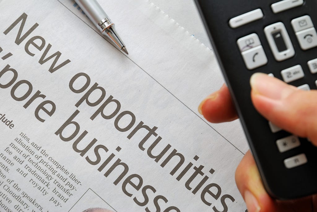 Closeup of new opportunities text on newspaper with hand dialing phone in foreground.