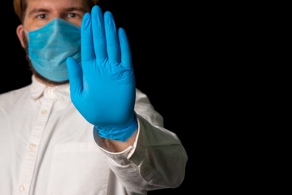 A photo of a medical personnel in a blue mask holding up a blue gloved hand.