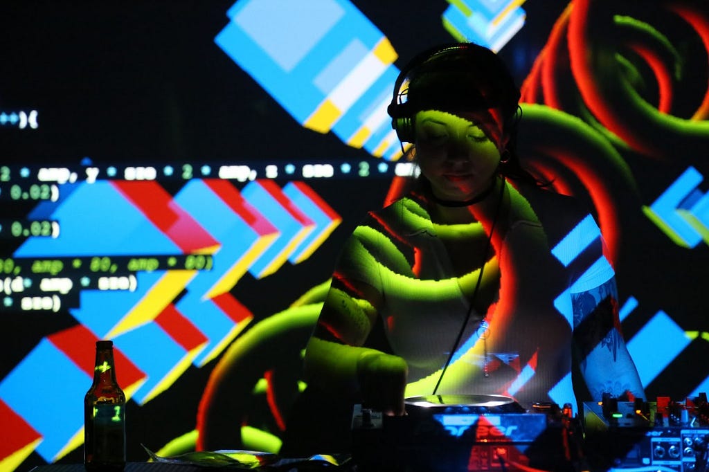 A person wearing headphones stands within a projection of p5.js code and visuals, in vivid blue, red, yellow, and black.