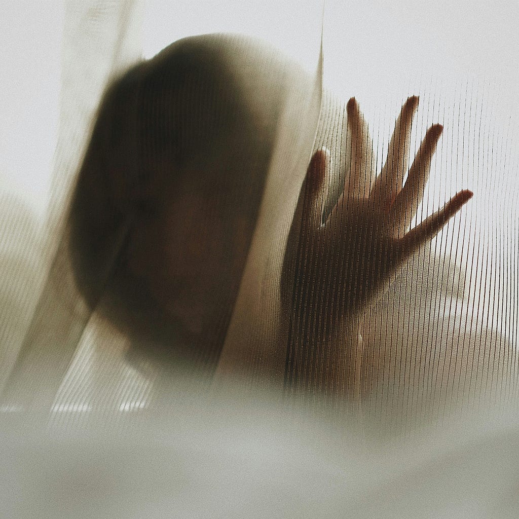 Sepia toned image of women with hand touching curtain, the image of her seen only shadow-like through the material.