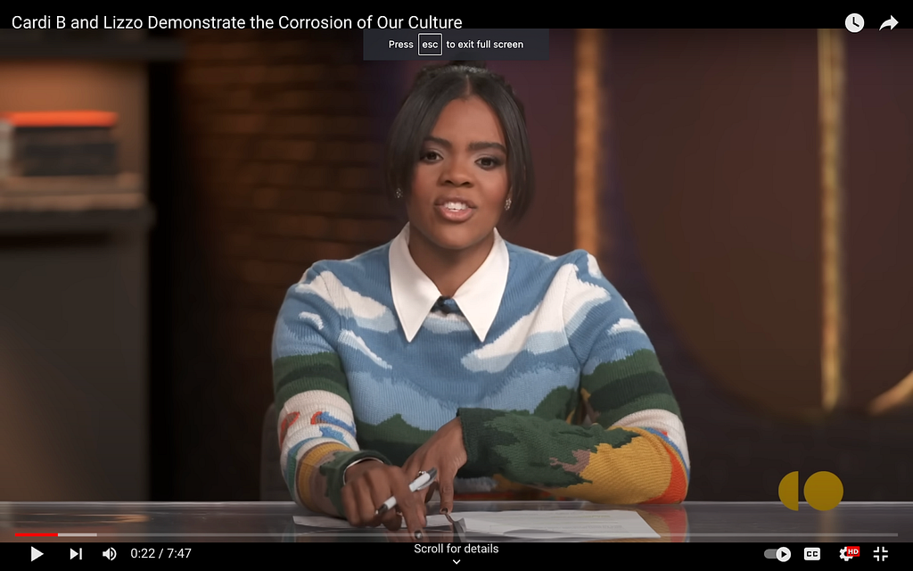 A screenshot from Candace Owens’s show on The Daily Wire. She wears a sweater with a brightly coloured landscape on it. There are green mountain tops, a blue sky and puffy white clouds. A long, pointed shirt collar from an item worn under the sweater is visible at the sweater’s neckline.