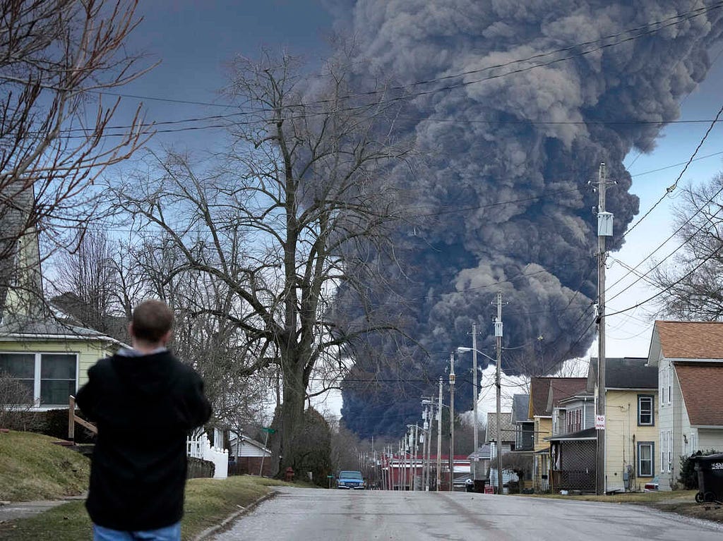 A plume of toxic smoke rises above East Palestine homes, an on-looker stands and watches