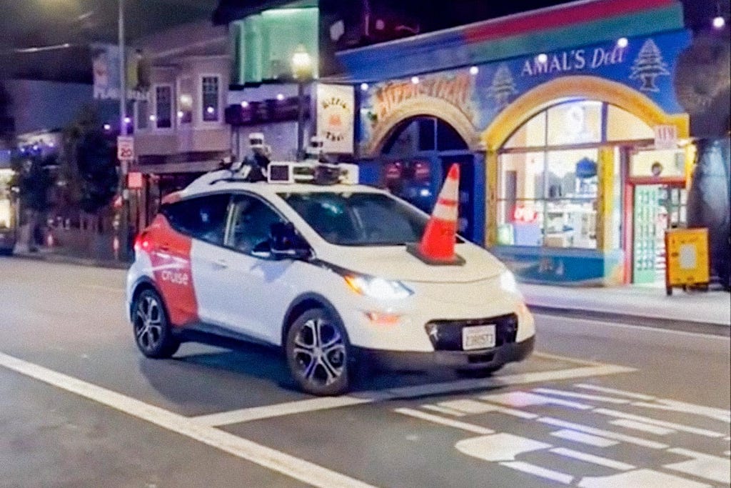 Image of a Waymo self-driving car on an urban street at nighttime, with a traffic cone on the bonnet.