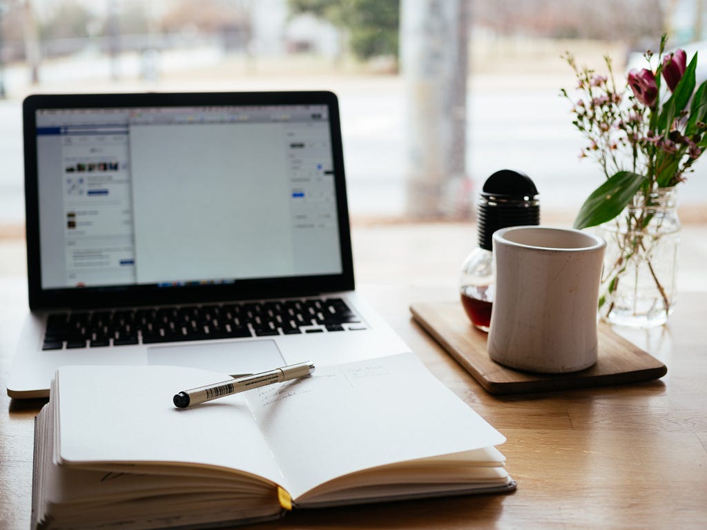 a desk with a notebook, pen, coffee cup and laptop