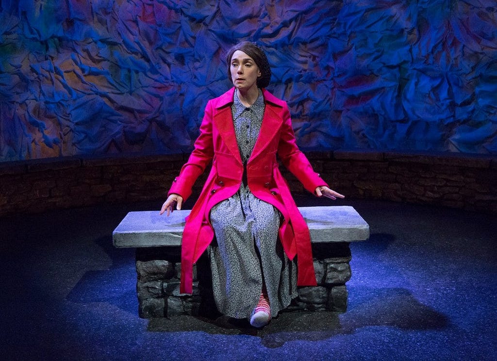 A woman with brown hair and a bright red coat over a dress sits on a bench with her eyes closed while she speaks