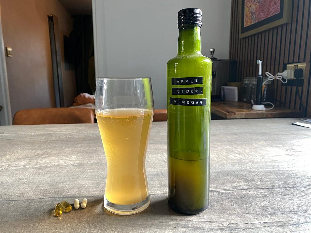 Apple cider vinegar in a bottle an a pint glass and some pills on a kitchen counter.