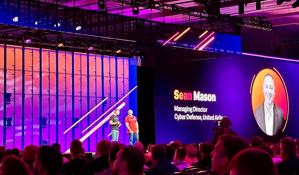 Two people are standing on a stage in front of an audience, with a large screen displaying the name Sean Mason and his professional title.
