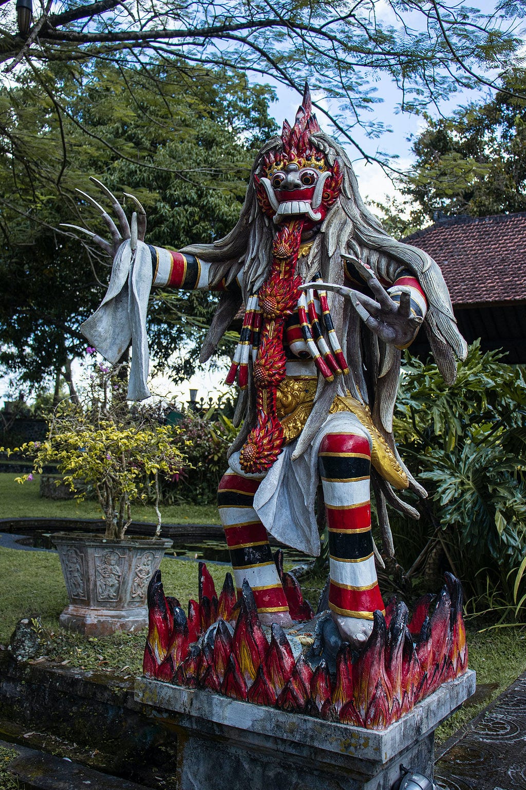 Statue of a Leyak in Bali, Indonesia, depicted as a mythological creature with a fearsome red and white mask featuring large bulging eyes and a protruding tongue adorned with a long, flowing gray mane. It stands with outstretched claws, wearing traditional Balinese striped garments, set upon a fiery-looking pedestal in a lush garden setting.