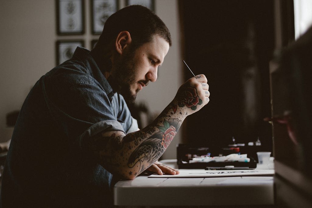 man with tribal flowers tattooed on his arm sits at a desk with paper on it and a pencil in his raised hand