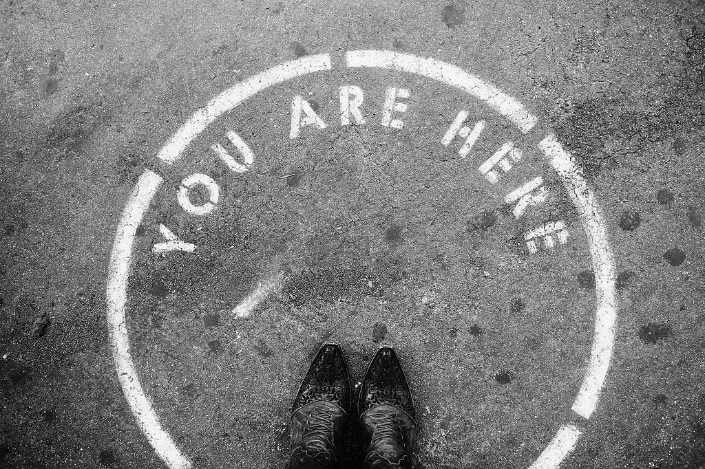A black and white photo of a person’s feet in fancy shoes standing in a circle painted on the floor, stating “you are here”.