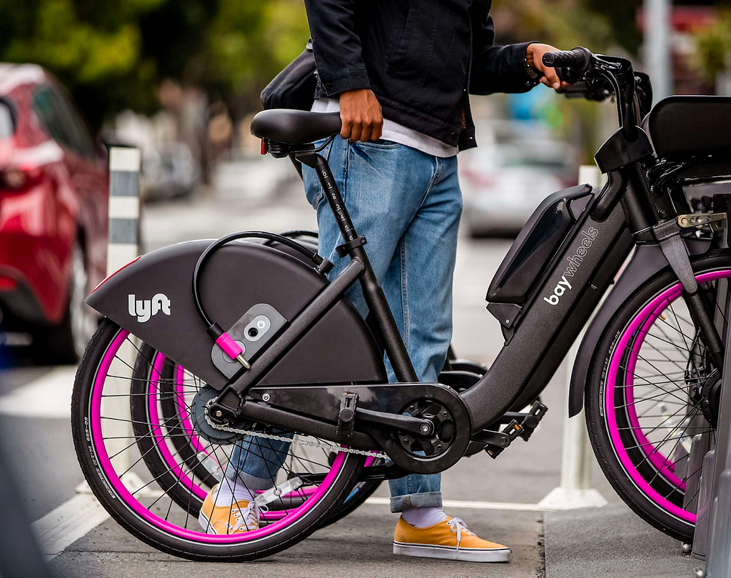 Person in jeans undocking a pink and black Lyft Bicycle from a dock on the side of the street