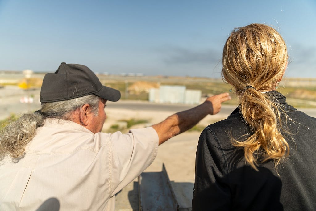 A man in a hat pointing toward the Gaza skyline, explaining the border crossing to a woman.