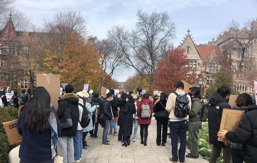 A group of people, viewed from the back, stand in front of university buildings. Several are holding cardboard signs, though the fronts of the signs are not visible.