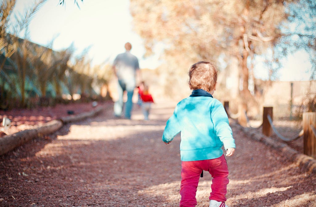 A child is following behind an adult who’s leading another child down a path.