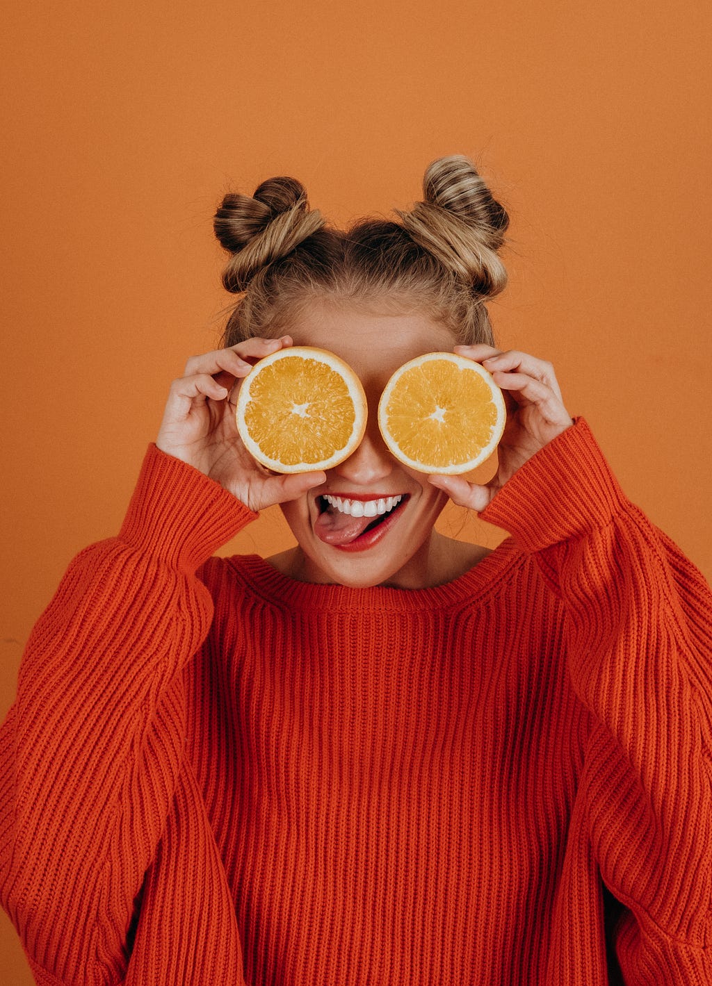 A young woman in a bright sweater smiling and holding two halves of orange in front of her eyes