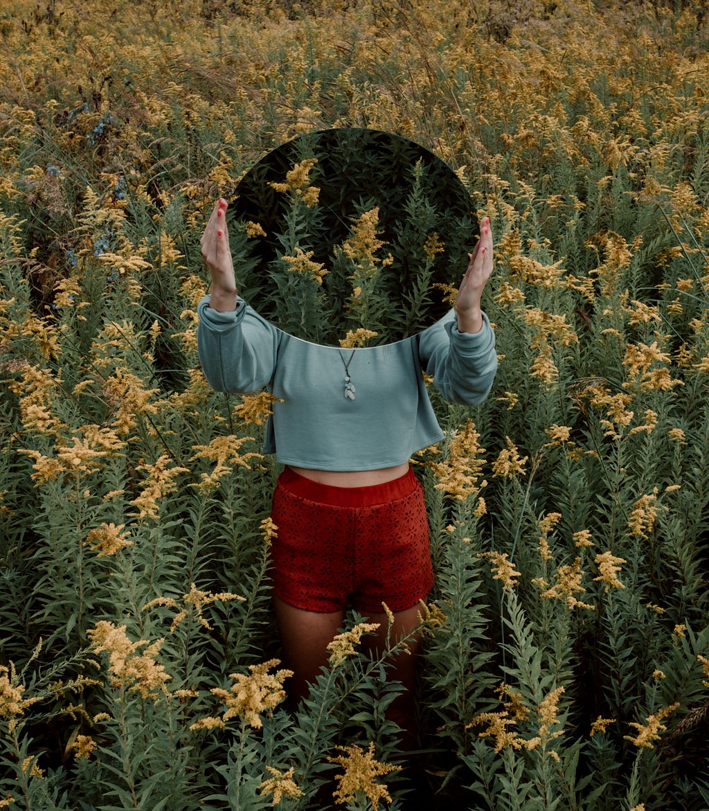 A girl holding a mirror over her face in a field of flowers