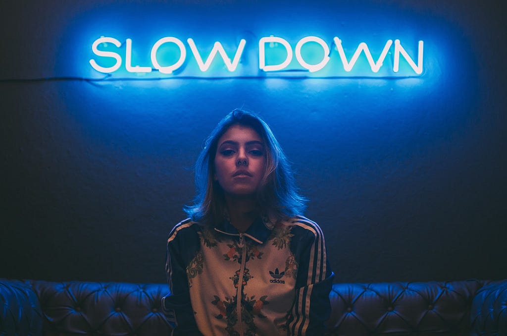 Picture of a woman sitting on a couch with a neon sign above her head saying slow down