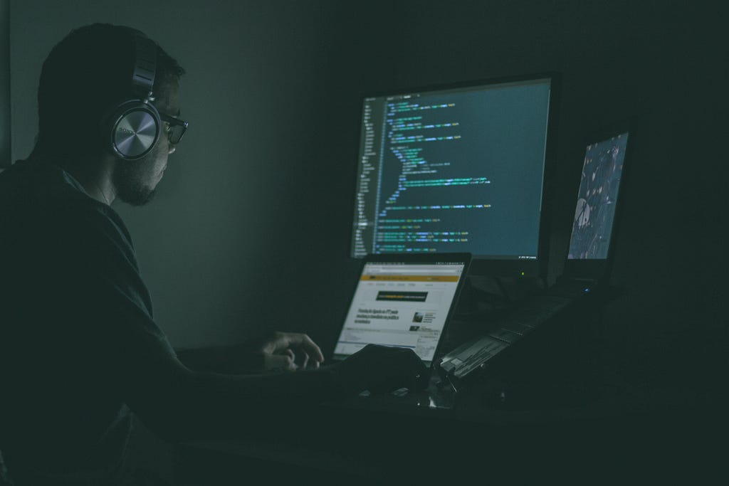 Photo of a man working in a dark room with a computer monitor and two laptop screens being the only light shining on the side of his face. He is wearing headphones, a form of computer coding can be seen on one of the screens.