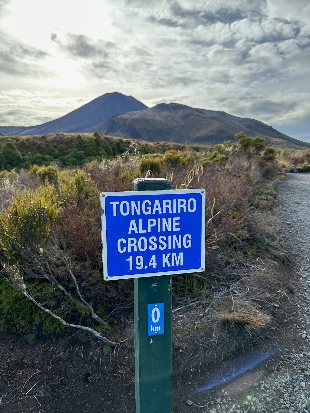 Volcano in distance. Trail leading to it with sign reading “Tongariro Alpine Crossing 19.4 km.”