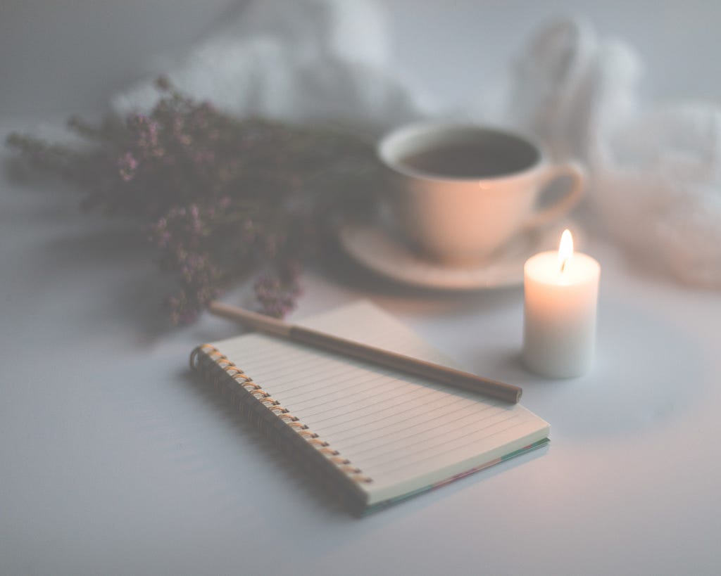 A blank notebook rests on a white background with what appears to be a wooden writing utensil atop its pages and a lit candle, a mug filled with dark liquid, and dried purple flowers arranged besides it.