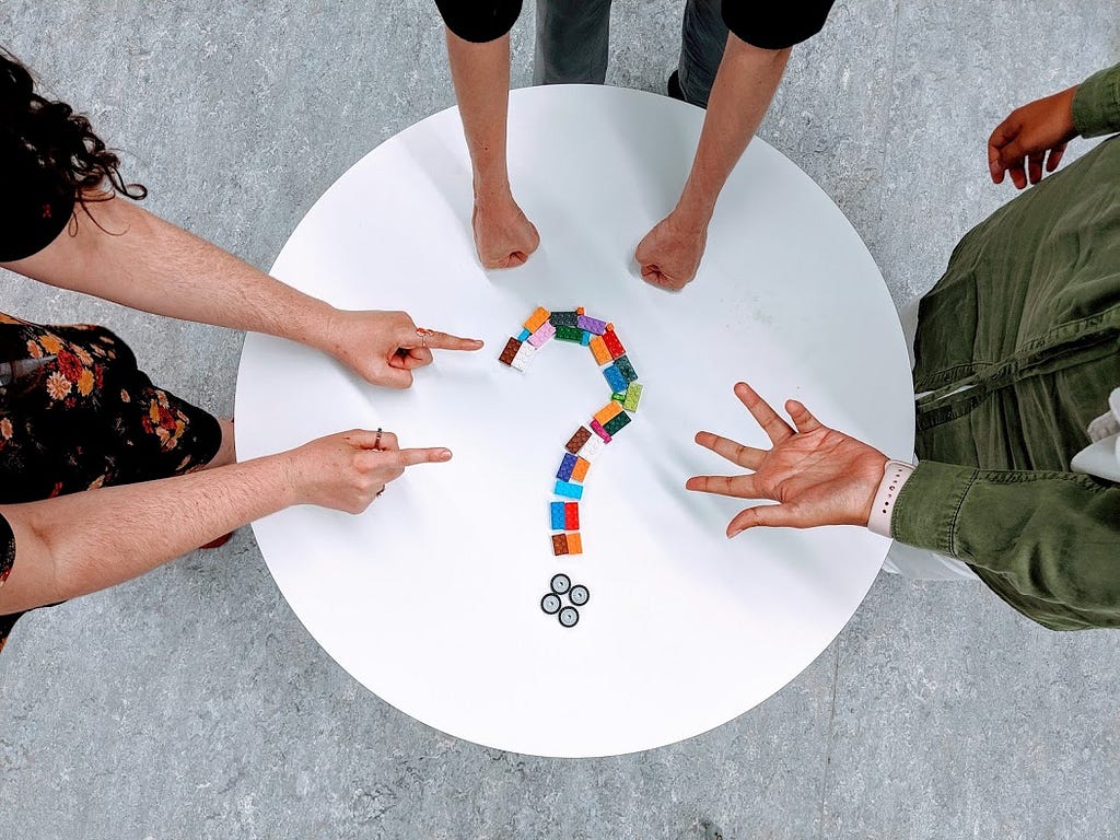 Three colleagues ask questions around a white, round table with a question mark made of lego placed the middle.