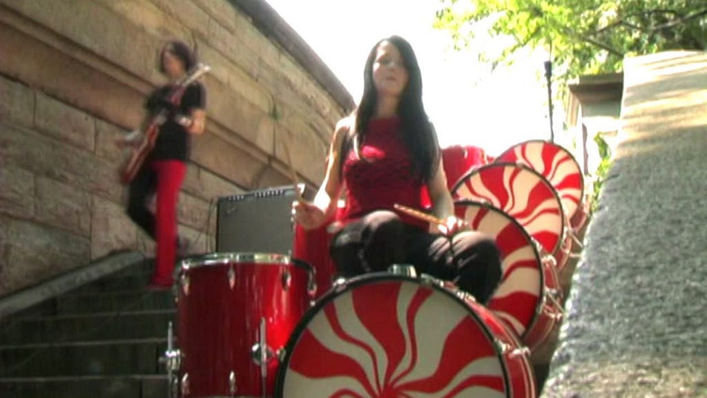 From The White Stripes’ “The Hardest Button to Button” music video; Jack White left walking down concrete park stairs playing guitar in black shirt and half black, half red pants — Meg White in a white tank top playing peppermint drums that have copied themselves on multiple steps