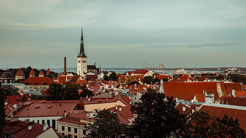 Many buildings with red roofs clustered around a central white tower with a gray sky and blue sea in the background