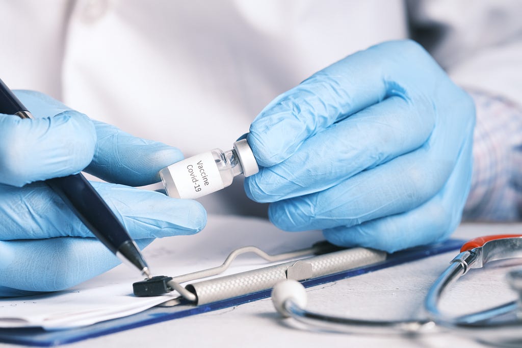 a pair of hands wearing blue nitrile gloves and holding a small jar labeled “Vaccine Covid-10” and a pen. in the background, the person’s body is shown partially wearing a labcoat, and in the foreground, one can see a clipboard and stethescope