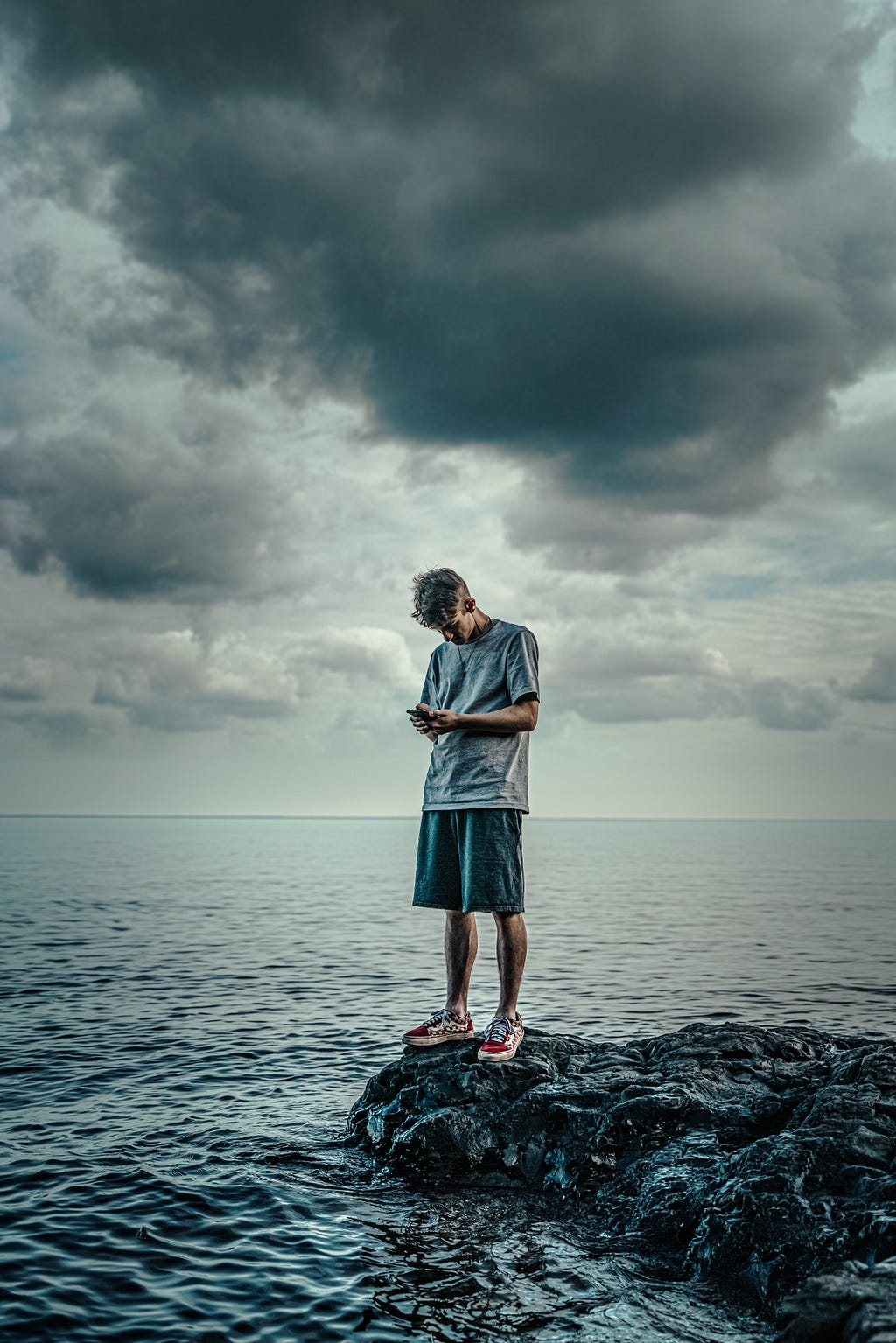 A photo of a man in a dark gray shirt, shorts, and red and white sneakers looking down at a cell phone in his hands. The day looks stormy and cloudy.