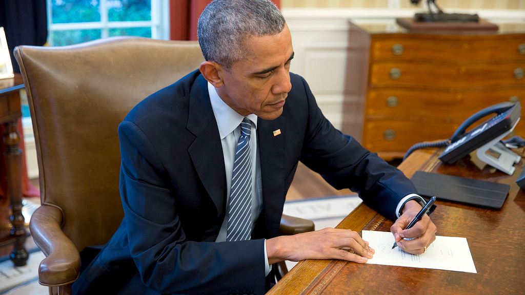 President Obama wearing a navy blue suit and striped tie, sitting at his desk hand writing a letter.