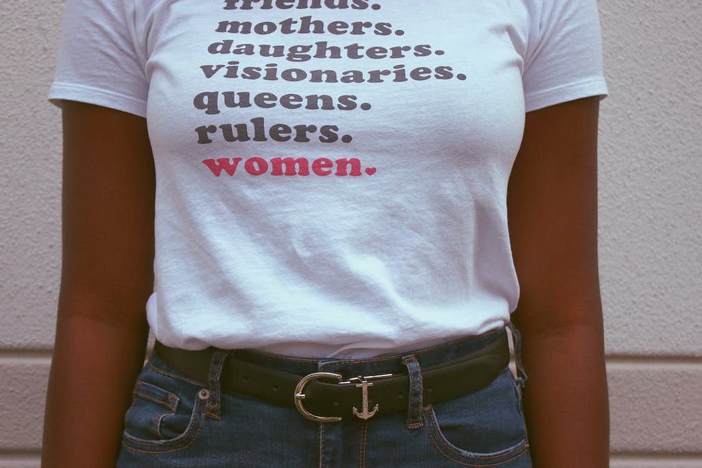 A close up of a woman wearing a t-shirt witht he slogan “friends, mothers, daughters, visionaries, queens, rulers, women.”