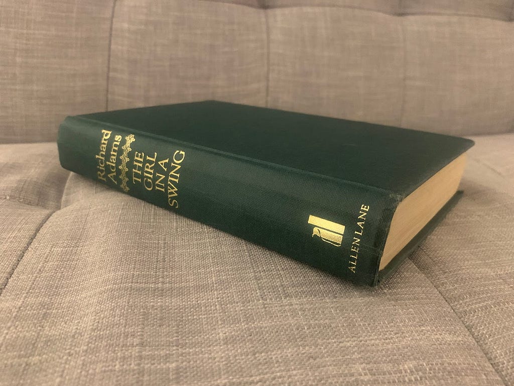 A third image of The Girl in a Swing by Richard Adams. This copy has no dust jacket and is laying on a grey couch. The hard cover is dark green, and the lettering on the spine is gold. It is a first-edition, second issue.