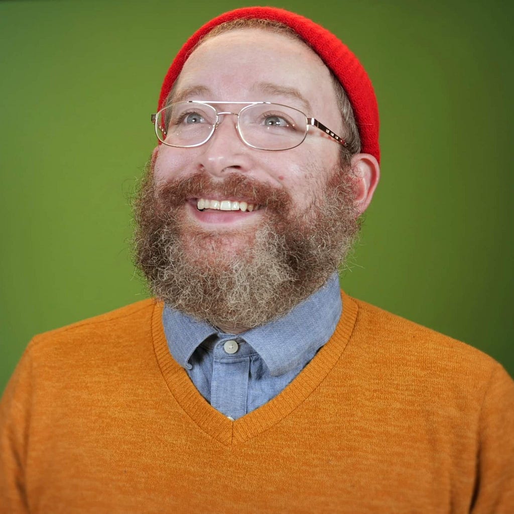 Photograph of Shawn Patrick, a white male with a large beard, smiling up and to the right of the camera. He wears thin golden-rimmed glasses, a buttoned-up blue collared shirt, a yellow sweater, and a bright red knitted cap. The background is a green screen.