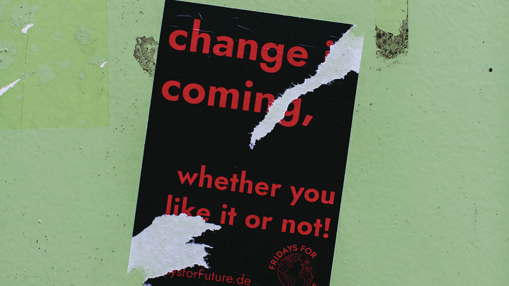 red and black sign with white tear marks, reading “change is coming whether you like it or not”
