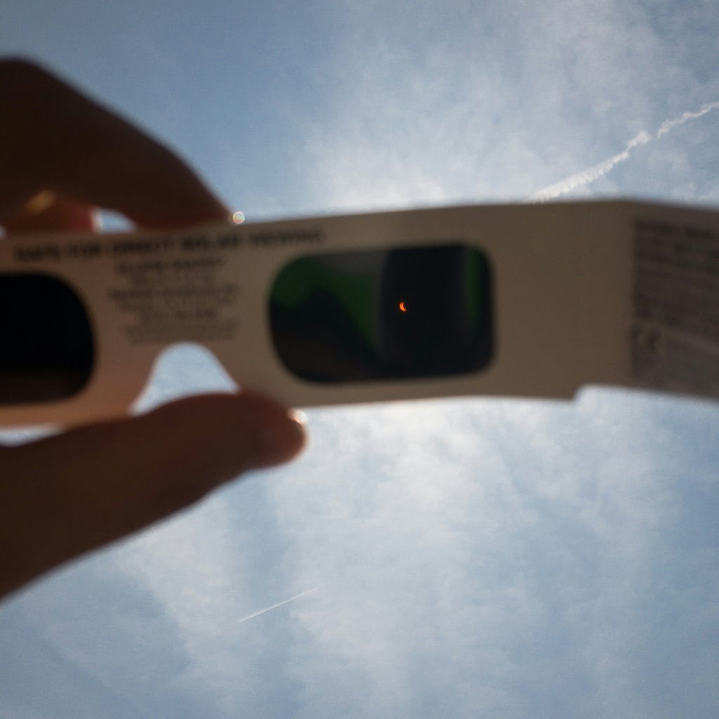 A photo from the perspective of someone lifting a pair of eclipse glasses to the sky. Through one of the dark lenses, we can see a speck of orange light — an eclipse as it is happening. The sky in the background is bright blue and mostly clear.