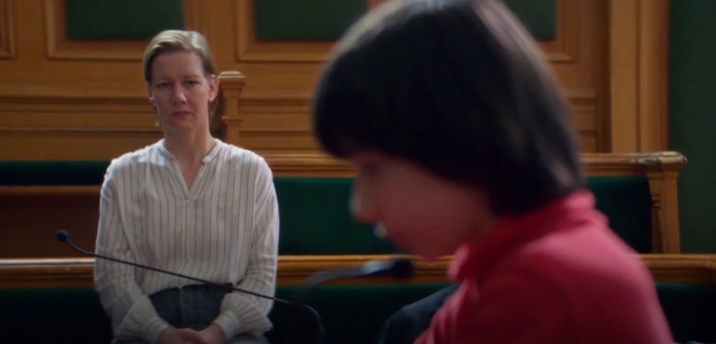 An image shows a snippet from the movie Anatomie d’une Chute. Sandra Huller as Sandra Voyter sits in court wearing a white top and black pants. Milo Machado-Graner, who plays Daniel in the film, is in the forefront.