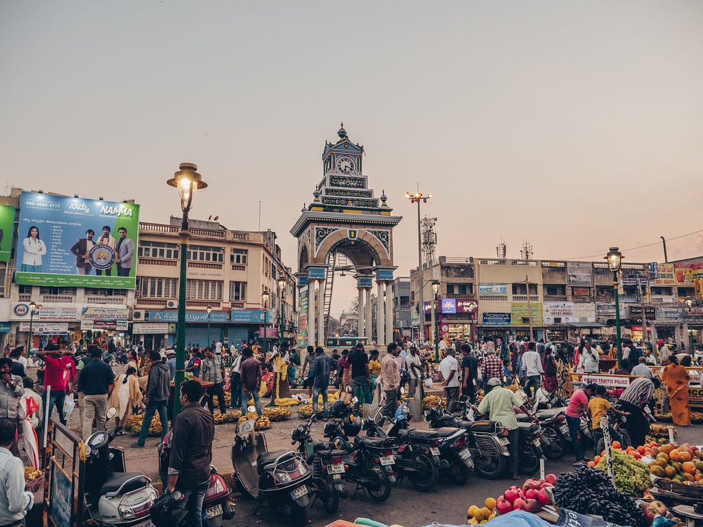 A crowded market in India at dusk, with people milling about, rows of parked motorbikes, and fresh produce on display.