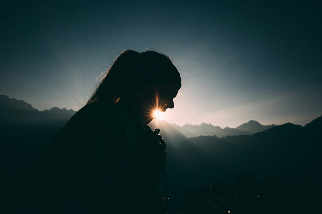 A girl’s silhouette against a backdrop pf mountains and a setting sun.