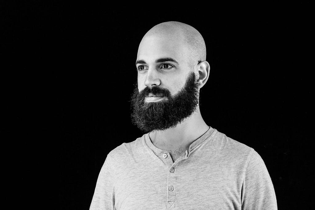 Photo by Peter Kolski photography. A headshot of Raphaël de Courville wearing a gray shirt against a black background. He is bald with a beard, smiling facing left.