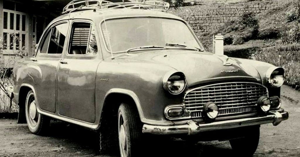 A vintage black-and-white photograph of a Hindustan Ambassador Mark I car, an iconic Indian automobile. The car has a rounded design, chrome grille, additional headlights, and a roof rack, parked in front of a house with a garden in the background.