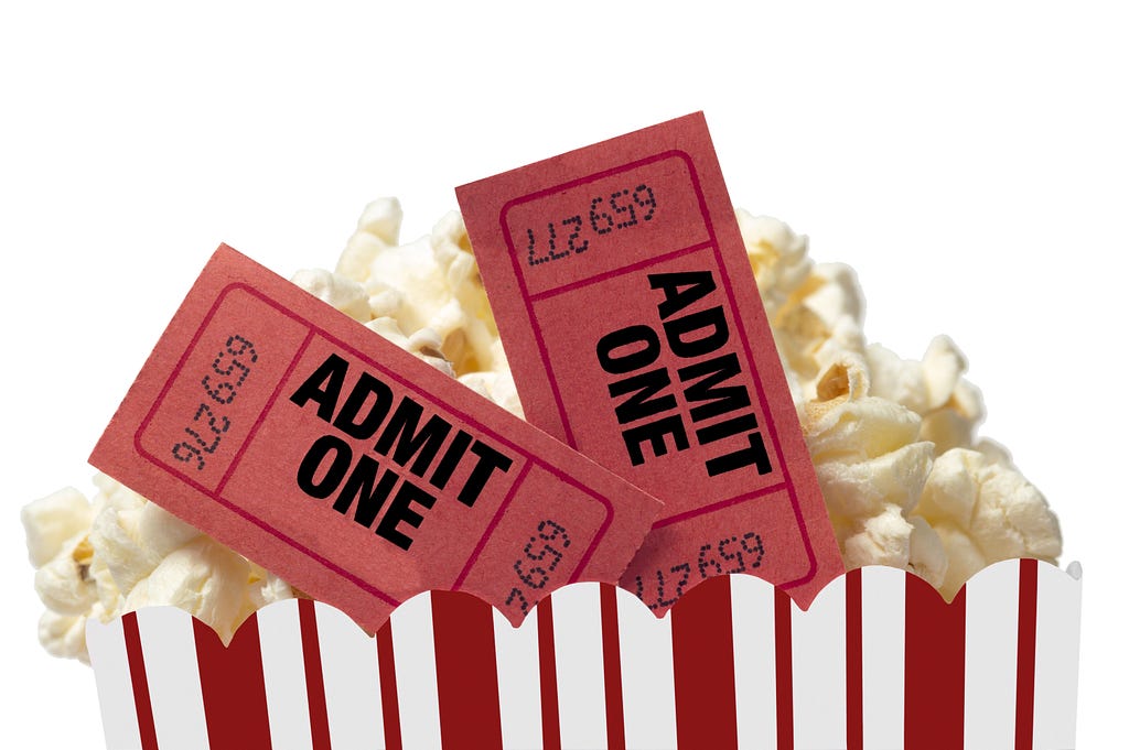 Two “Admit One” movie tickets are tucked into the top of a paper bucket of movie popcorn.