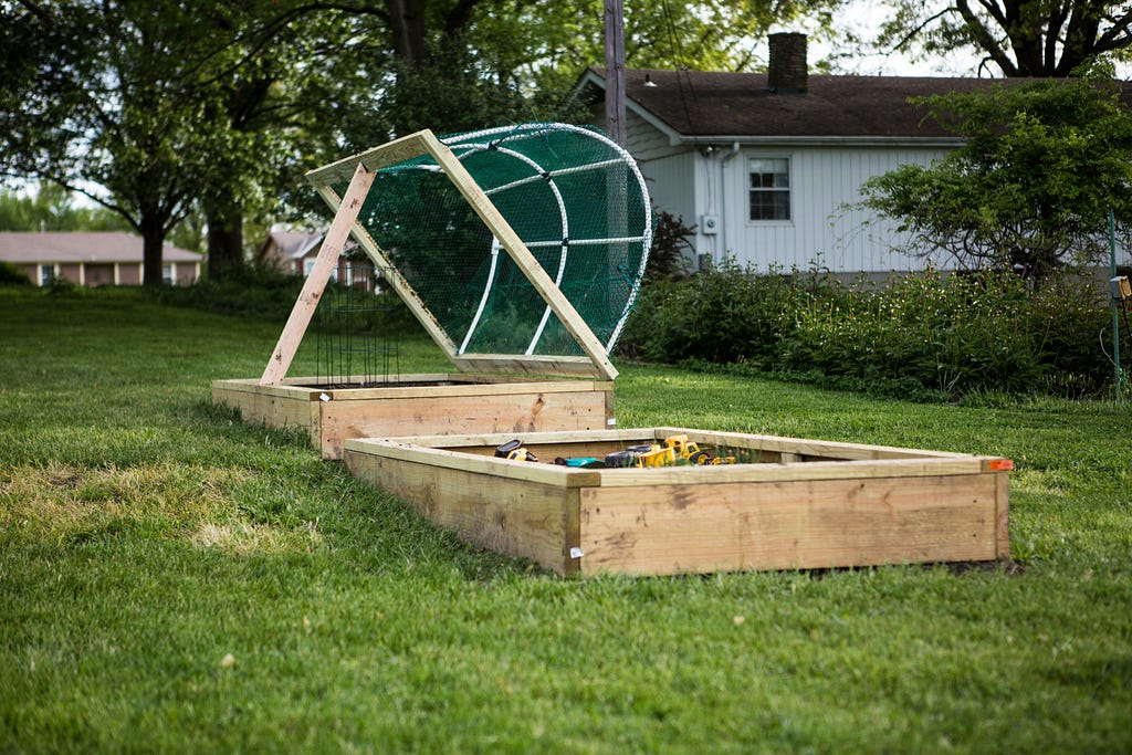 Two raised garden beds in the middle of a lawn with a house in the background. One bed is open but has a glass covering that can go over it